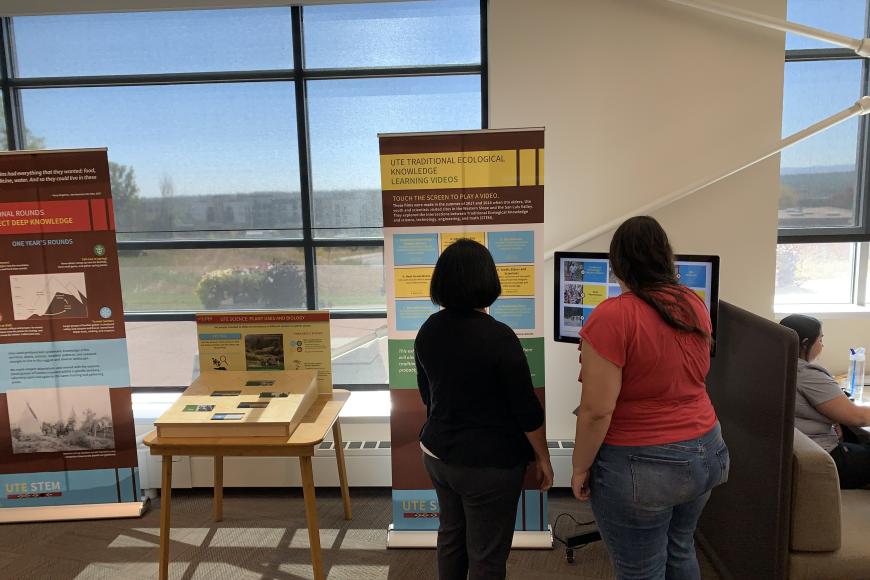 Two women reading a pop-up panel about Ute knowledge, in front of a large window.