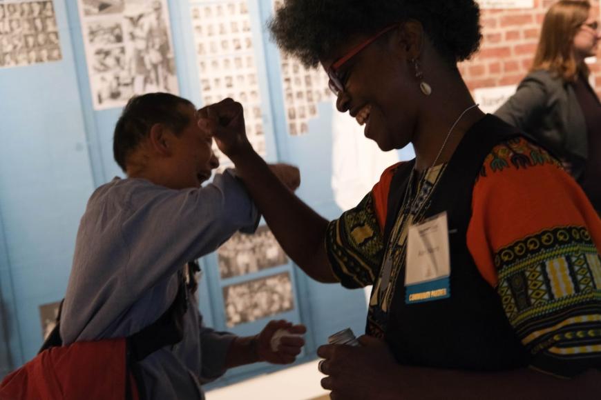 Adri Norris, a Black woman, and a man of asian descent dance and celebrate with their fists raised in front of a colorful mural.