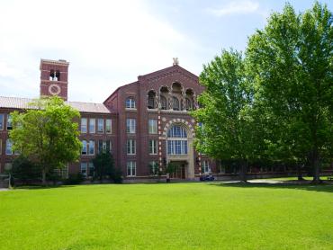 View of a red brick front-gabled portion of a building, with a clock tower visible in the background and green trees and lawn.