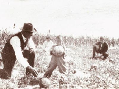 A Black family, including two adult men and a small child kneel in a field. The child holds a large vegetable and is smiling for the camera.