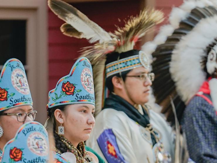 A group of young women and a tribal leader from the Ute tribe stand in the History Colorado Center in traditional garb.