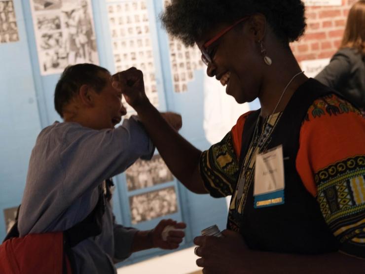Adri Norris, a Black woman, and a man of asian descent dance and celebrate with their fists raised in front of a colorful mural.