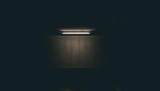 Photo of one single long flourescent light bulb, mounted to the ceiling of a dark area such as an empty parking garage. The light is illuminated, casting a glow over one concrete wall. The rest of the area remains dark.