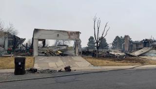 A burned house sits on a street. Only one front wall is left partially standing, and through the gaping garage door a pile of rubble is visible.