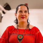 Photo of Adrianna Abarca, standing in front of a white background. She has long black hair that is pulled tightly to the back of her head, with dark eyes and red lips, she is smiling. She is wearing a bright red blouse and a beaded medallion necklace.