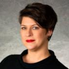 Upclose photo of Ann Sneesby-Koch. She has short dark hair that is parted on her right side and sweeps to the left. She is smiling. She is wearing a black cowl neck blouse, and red lipstick, with black earrings.