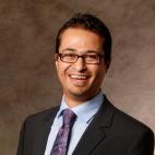Photo of a man wearing a light blue dress shirt, purple and blue paisley necktie, and dark colored suit jacket. He is smiling. He has short cut dark hair and a small patch of facial hair on his chin. He is smiling, and he wears rectangular shaped eyewear.