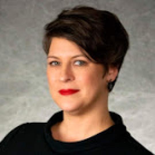 Ann Sneesby-Koch Upclose photo of Ann Sneesby-Koch. She has short dark hair that is parted on her right side and sweeps to the left. She is smiling. She is wearing a black cowl neck blouse, and red lipstick, with black earrings.