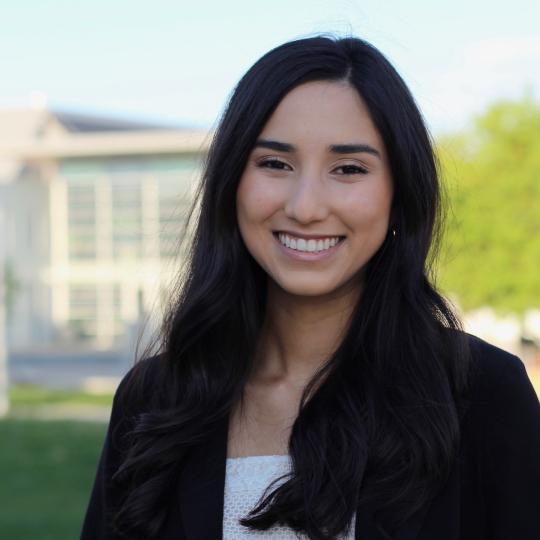 Photo of Raena Vigil, standing outdoors. Grass, trees, and a large building are in the distance behind.
